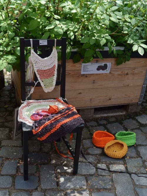 Auf einem Stuhl und auf dem Pflaster daneben sind Handarbeitsst&uuml;cke ausgestellt. Der Stuhl steht vor einem Hochbeet mit &uuml;ppigem Gr&uuml;nzeug.