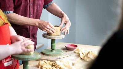Ein Kind und eine erwachsene Person stehen vor einem Tisch. Darauf stehen zwei St&auml;nder, 2 T&uuml;cher, eine Schale mit Wasser und ein T&ouml;pferwerkzeug. Auf den beiden St&auml;nder werden Ton St&uuml;cke geformt
