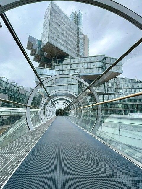 Ein futuristischer Glasgang, der zu einem modernen Geb&auml;ude mit auff&auml;lliger Architektur f&uuml;hrt.