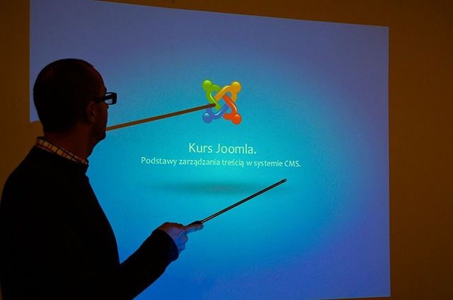 Eine Person zeigt mit einem Zeigestab auf eine projizierte Pr&auml;sentation mit dem Joomla-Logo. Der Text lautet: "Kurs Joomla. Grundlagen der Inhaltsverwaltung im CMS