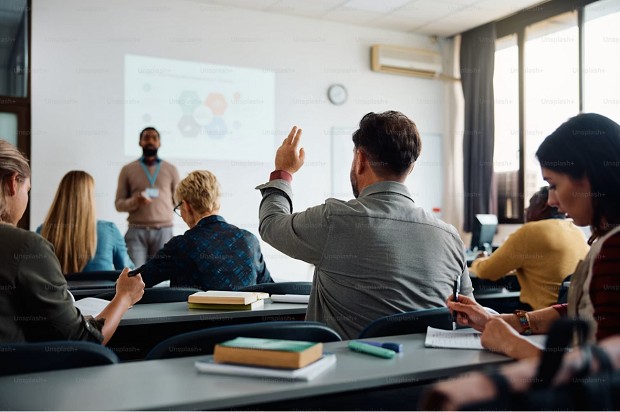 Klassenraum mit Lehrkraft vorn und ein Teilnehmer meldet sich