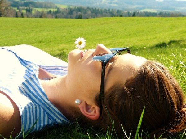 Eine Frau liegt entspannt auf einer Wiese im Sonnenschein und hat ein G&auml;nsebl&uuml;mchen zwischen den Lippen.
